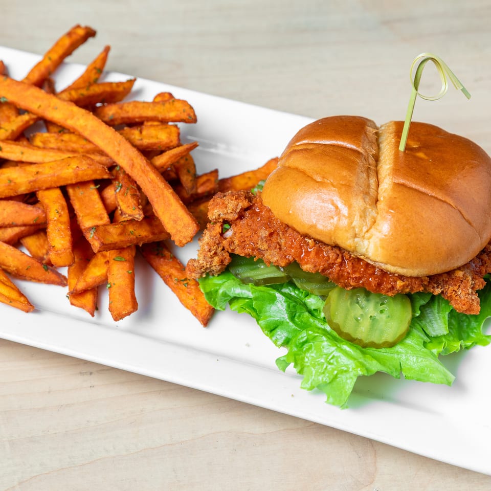 Crispy fried chicken sandwich with lettuce and pickles served alongside seasoned sweet potato fries on a white plate.