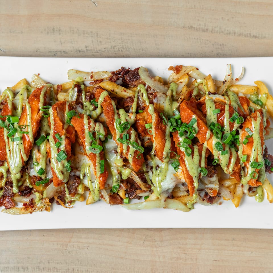 Loaded fries topped with crispy chicken, bacon, melted cheese, onions, green sauce, hot sauce, and scallions on a white platter.
