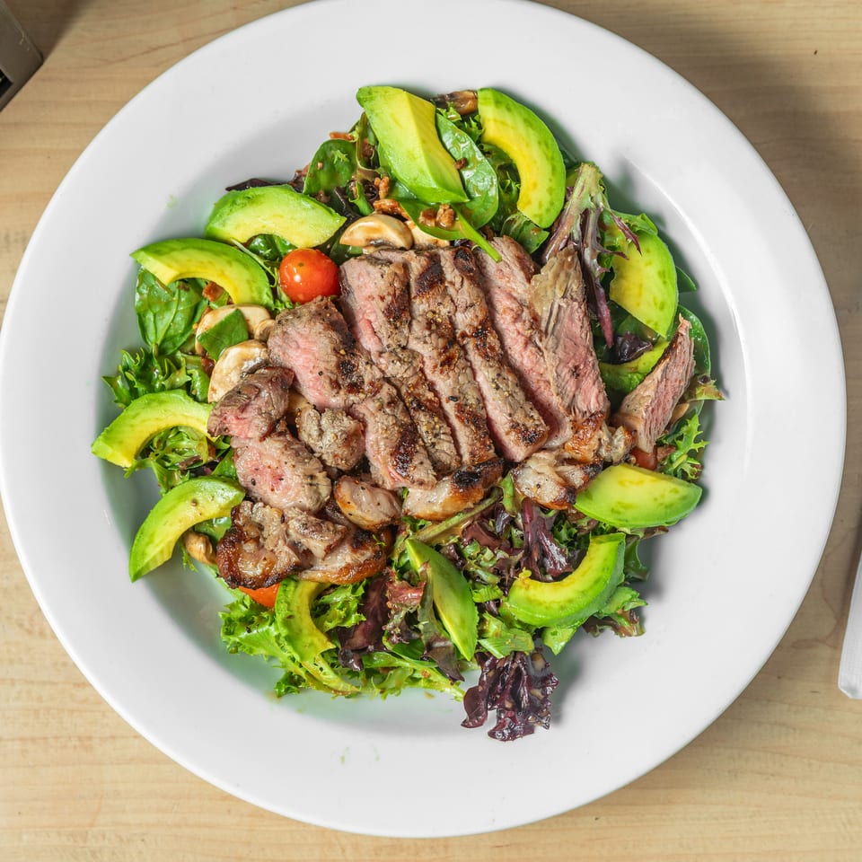 Sliced grilled steak atop mixed greens with avocado, mushrooms, cherry tomatoes, and crispy bacon on a white plate.