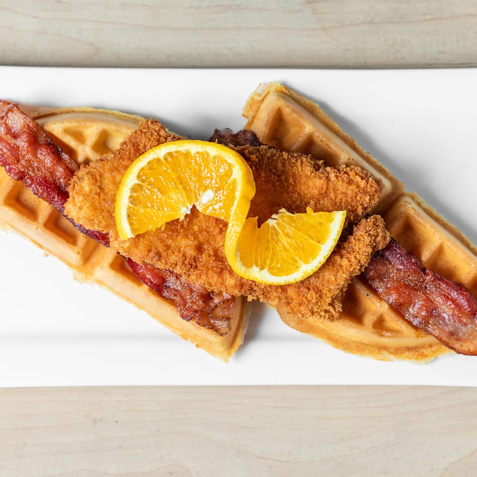 Crispy fried chicken, bacon, and orange slices arranged on golden waffles atop a white plate.